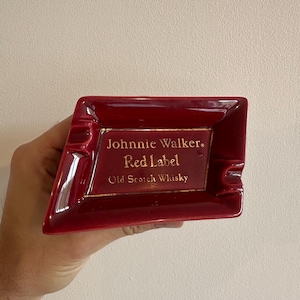 May include: A red ceramic ashtray with the text "Johnnie Walker Red Label Old Scotch Whisky" in gold lettering. The ashtray has a rectangular shape with a beveled edge and a small indentation for resting cigarettes.