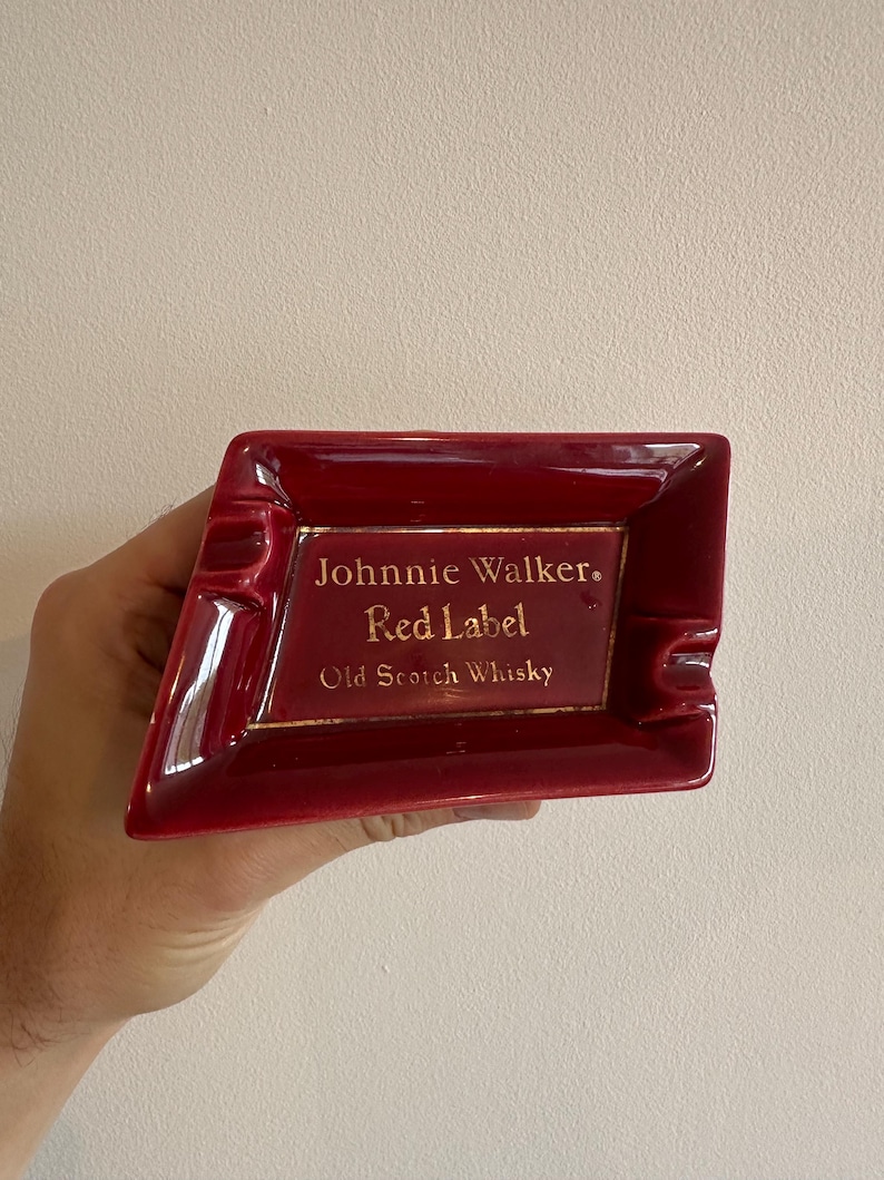 May include: A red ceramic ashtray with the text "Johnnie Walker Red Label Old Scotch Whisky" in gold lettering. The ashtray has a rectangular shape with a beveled edge and a small indentation for resting cigarettes.