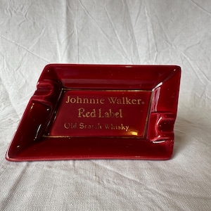 May include: A red ceramic ashtray with a rectangular shape and a slanted side. The ashtray features the text "Johnnie Walker Red Label Old Scotch Whisky" in gold lettering. The ashtray has two built-in rests for cigarettes.