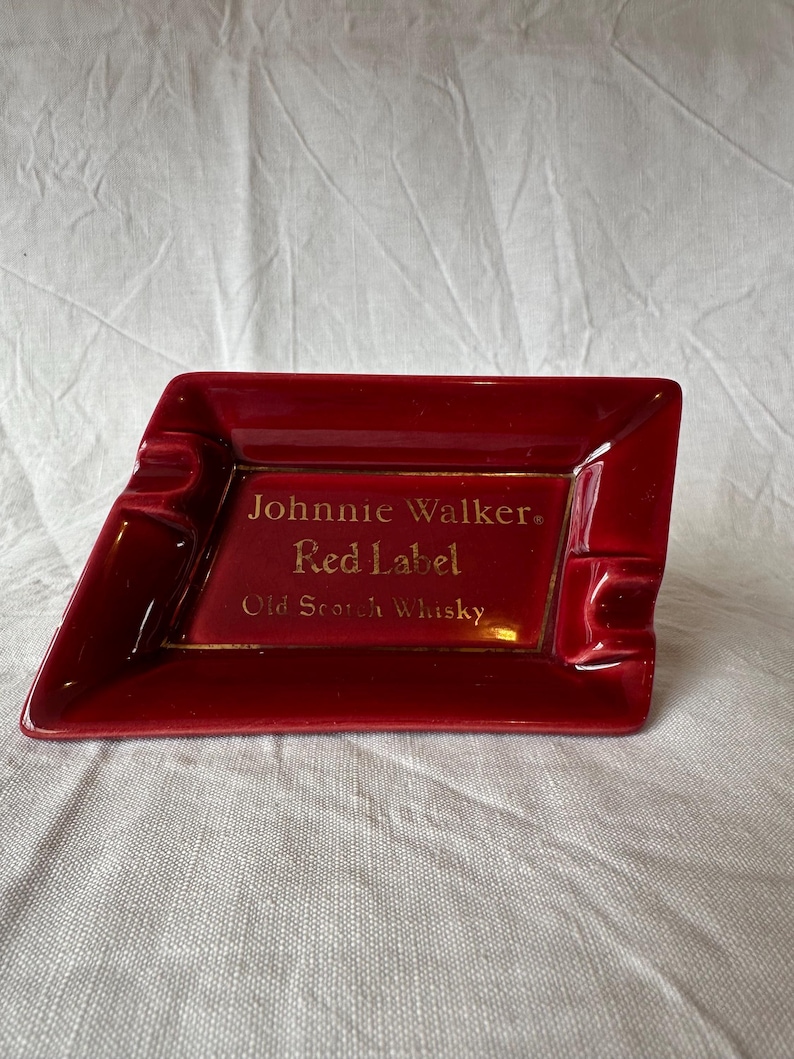 May include: A red ceramic ashtray with a rectangular shape and a slanted side. The ashtray features the text "Johnnie Walker Red Label Old Scotch Whisky" in gold lettering. The ashtray has two built-in rests for cigarettes.