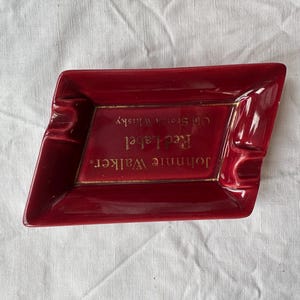 May include: A red ceramic ashtray with a diamond shape. The ashtray has a gold border and features the text "Johnnie Walker Red Label Old Scotch Whisky."