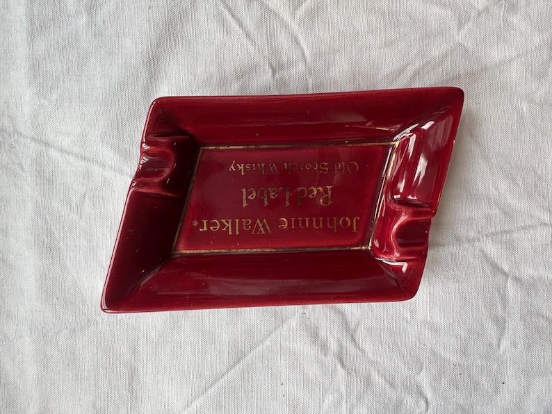 May include: A red ceramic ashtray with a diamond shape. The ashtray has a gold border and features the text "Johnnie Walker Red Label Old Scotch Whisky."