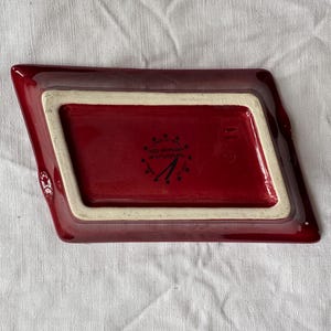 May include: A burgundy-colored ceramic dish with a rectangular interior and angled sides. The interior is a darker shade of burgundy, framed by a cream-colored border. The underside features a logo and text.