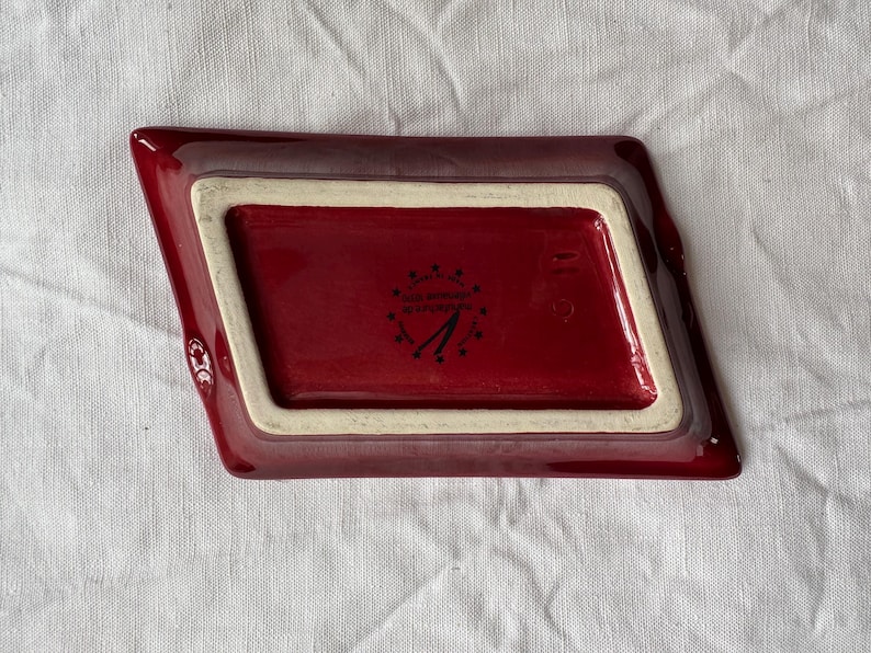 May include: A burgundy-colored ceramic dish with a rectangular interior and angled sides. The interior is a darker shade of burgundy, framed by a cream-colored border. The underside features a logo and text.
