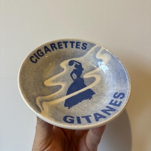 Puede incluir: Un cenicero pequeño y redondo con un diseño moteado azul y blanco. Las palabras "CIGARETTES GITANES" están impresas en azul alrededor del borde. Una silueta azul de una bailarina está en el centro.