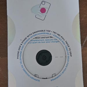 May include: A PopSockets PopGrip on a white and grey card. The PopGrip is black and circular. The card has text that says "POPSOCKETS" and "POPGRIP". It also has instructions and a diagram of a phone.