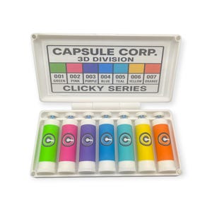 Op de afbeelding: Een witte plastic doos met zeven kleurrijke Clicky Series markers. Elke marker heeft een andere kleur: groen, roze, paars, blauw, turquoise, geel en oranje. De doos heeft de tekst "CAPSULE CORP. 3D DIVISION" en een kleurenkaart.