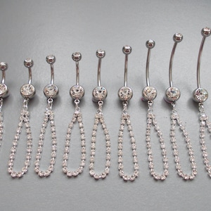 May include: A selection of silver belly button rings, each with a dangling teardrop-shaped chain of crystals. The rings have a clear, round gem at the top and a larger gem at the centre. The crystals shimmer against a grey backdrop.