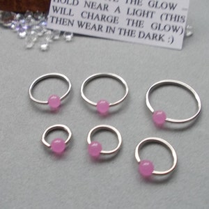 Puede incluir: Seis aros de nariz plateados con una peque&ntilde;a cuenta redonda de color rosa en el centro. Los aros var&iacute;an en tama&ntilde;o y est&aacute;n dispuestos sobre una superficie gris. Una peque&ntilde;a tarjeta blanca con texto negro dice: "SOSTENER CERCA DE UNA LUZ (ESTO CARGAR&Aacute; EL BRILLO) LUEGO USAR EN LA OSCURIDAD :)"