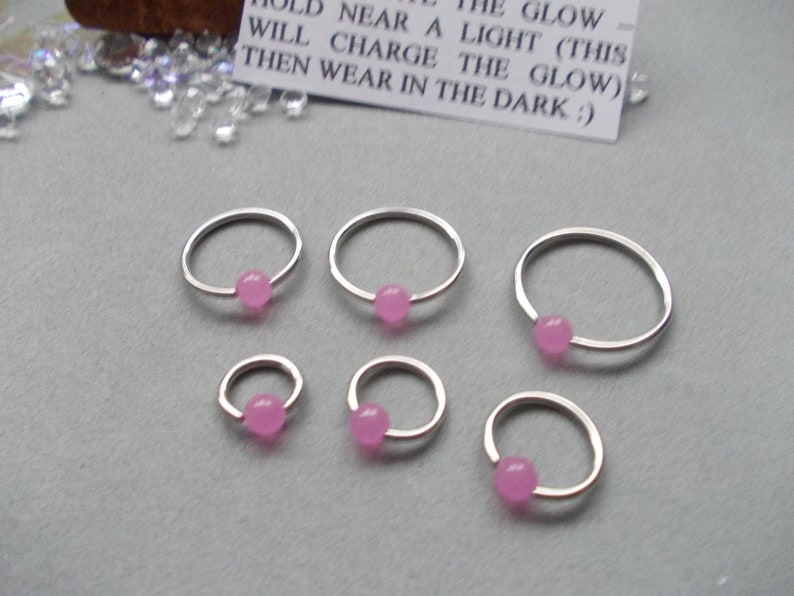 Puede incluir: Seis aros de nariz plateados con una peque&ntilde;a cuenta redonda de color rosa en el centro. Los aros var&iacute;an en tama&ntilde;o y est&aacute;n dispuestos sobre una superficie gris. Una peque&ntilde;a tarjeta blanca con texto negro dice: "SOSTENER CERCA DE UNA LUZ (ESTO CARGAR&Aacute; EL BRILLO) LUEGO USAR EN LA OSCURIDAD :)"