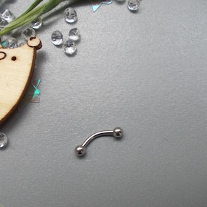 May include: A curved eyebrow piercing made of silver-colored metal. The jewelry has a small ball on each end. The piercing is on a gray surface, with decorative elements in the background.