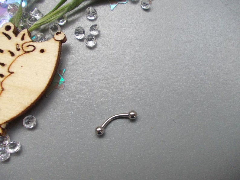 May include: A curved eyebrow piercing made of silver-colored metal. The jewelry has a small ball on each end. The piercing is on a gray surface, with decorative elements in the background.