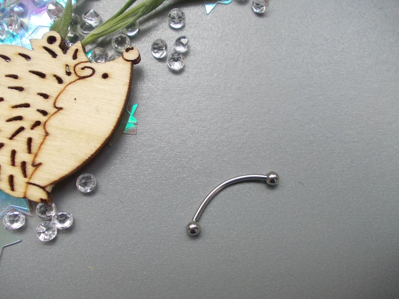 May include: A curved eyebrow piercing with two small silver balls on each end. The jewelry is displayed on a gray surface with a wooden hedgehog cutout, clear rhinestones, and iridescent confetti.