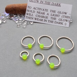 Puede incluir: Seis pendientes de aro dorados con una cuenta verde ne&oacute;n en el centro. Una peque&ntilde;a tarjeta con instrucciones sobre c&oacute;mo activar la funci&oacute;n que brilla en la oscuridad es visible. El texto de la tarjeta dice: "GLOW IN THE DARK. TO ACTIVATE THE GLOW - HOLD NEAR A LIGHT (THIS WILL CHARGE THE GLOW) THEN WEAR IN THE DARK :)"