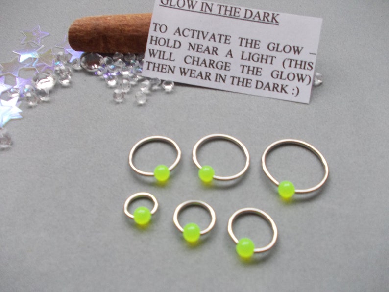Puede incluir: Seis pendientes de aro dorados con una cuenta verde ne&oacute;n en el centro. Una peque&ntilde;a tarjeta con instrucciones sobre c&oacute;mo activar la funci&oacute;n que brilla en la oscuridad es visible. El texto de la tarjeta dice: "GLOW IN THE DARK. TO ACTIVATE THE GLOW - HOLD NEAR A LIGHT (THIS WILL CHARGE THE GLOW) THEN WEAR IN THE DARK :)"