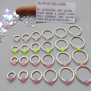 Puede incluir: Una variedad de pendientes de aro plateados con cuentas que brillan en la oscuridad en varios colores, incluyendo blanco, verde ne&oacute;n, rosa y morado. La imagen incluye el texto "GLOW IN THE DARK".