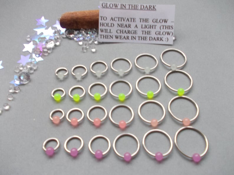 Puede incluir: Una variedad de pendientes de aro plateados con cuentas que brillan en la oscuridad en varios colores, incluyendo blanco, verde ne&oacute;n, rosa y morado. La imagen incluye el texto "GLOW IN THE DARK".