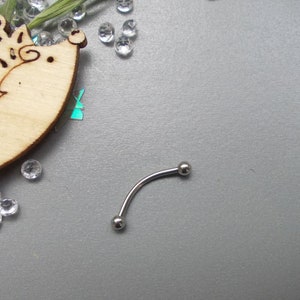 May include: A curved, silver-colored eyebrow piercing barbell with two small, round balls at each end. The jewelry is displayed on a gray surface, with a wooden hedgehog cutout and scattered clear rhinestones in the background.