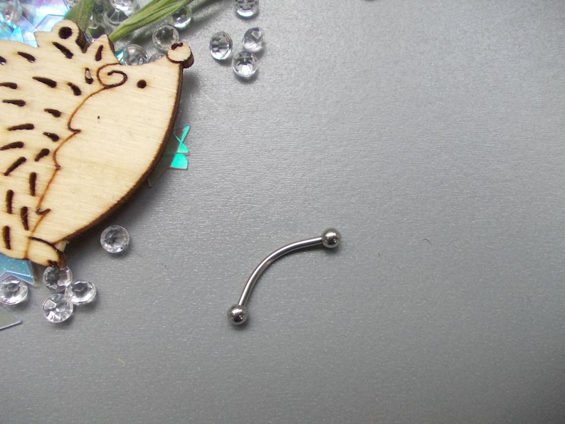 May include: A curved, silver-colored eyebrow piercing barbell with two small, round balls at each end. The jewelry is displayed on a gray surface, with a wooden hedgehog cutout and scattered clear rhinestones in the background.