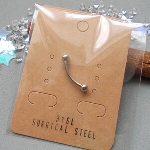 May include: A curved eyebrow piercing made of surgical steel, packaged on a brown card. The card has the text "316L SURGICAL STEEL" printed on it. The piercing is silver and has a small ball on each end.
