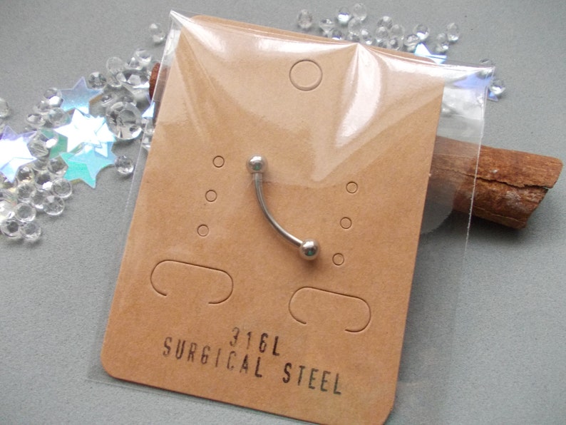 May include: A curved eyebrow piercing made of surgical steel, packaged on a brown card. The card has the text "316L SURGICAL STEEL" printed on it. The piercing is silver and has a small ball on each end.