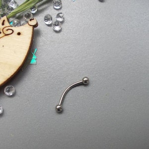 May include: A curved, silver-colored eyebrow piercing with two small, round balls at each end. The jewelry is set against a gray background, with decorative clear crystals and a wooden hedgehog cutout.