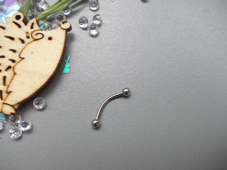 May include: A curved, silver-colored eyebrow piercing with two small, round balls at each end. The jewelry is set against a gray background, with decorative clear crystals and a wooden hedgehog cutout.