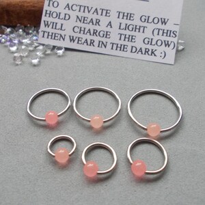 Puede incluir: Seis aros de nariz plateados con una peque&ntilde;a cuenta rosa claro. Una peque&ntilde;a tarjeta blanca con texto negro dice: "PARA ACTIVAR EL BRILLO - SOSTENER CERCA DE UNA LUZ (ESTO CARGAR&Aacute; EL BRILLO) LUEGO USAR EN LA OSCURIDAD :)"