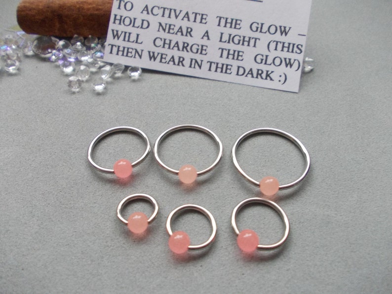 Puede incluir: Seis aros de nariz plateados con una peque&ntilde;a cuenta rosa claro. Una peque&ntilde;a tarjeta blanca con texto negro dice: "PARA ACTIVAR EL BRILLO - SOSTENER CERCA DE UNA LUZ (ESTO CARGAR&Aacute; EL BRILLO) LUEGO USAR EN LA OSCURIDAD :)"