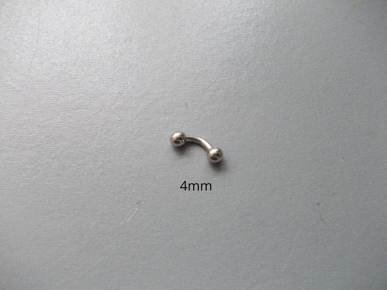 May include: A curved, silver-colored eyebrow piercing with two small, round balls at each end. The text "4mm" is printed below the piercing, indicating its size. The jewelry is set against a gray background.