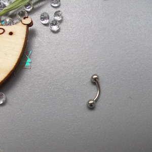 May include: A curved, silver-colored eyebrow piercing barbell with two small, round balls at each end. The jewelry is displayed on a textured, gray surface, with decorative elements in the background.