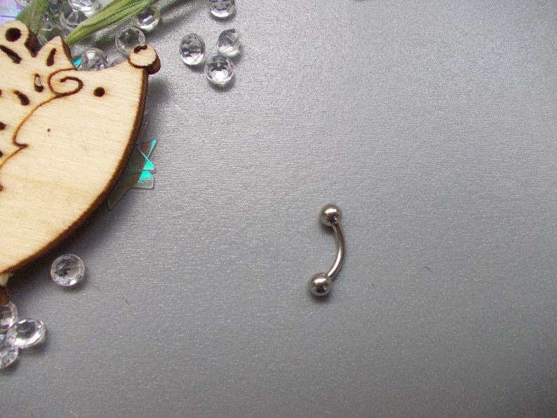May include: A curved, silver-colored eyebrow piercing barbell with two small, round balls at each end. The jewelry is displayed on a textured, gray surface, with decorative elements in the background.