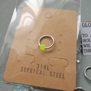 Puede incluir: Un aro de nariz plateado con una cuenta verde ne&oacute;n, exhibido en una tarjeta marr&oacute;n. La tarjeta est&aacute; etiquetada como "316L SURGICAL STEEL". El anillo est&aacute; empaquetado en una bolsa de pl&aacute;stico transparente. Una peque&ntilde;a tarjeta dice "GLOW IN THE..." con instrucciones.