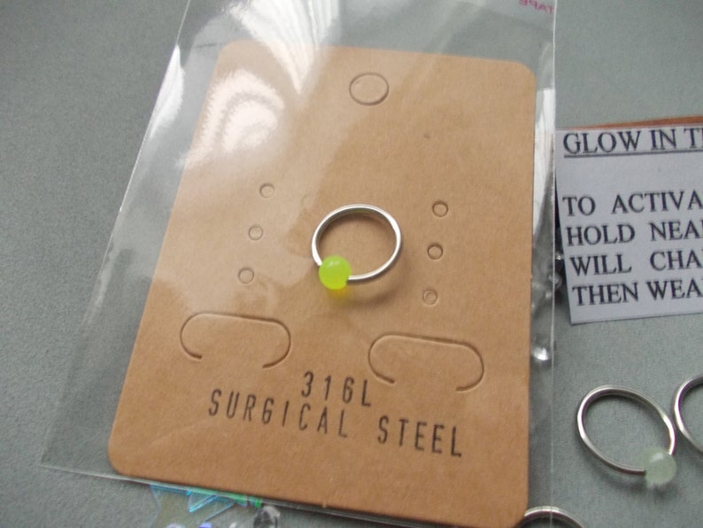 Puede incluir: Un aro de nariz plateado con una cuenta verde ne&oacute;n, exhibido en una tarjeta marr&oacute;n. La tarjeta est&aacute; etiquetada como "316L SURGICAL STEEL". El anillo est&aacute; empaquetado en una bolsa de pl&aacute;stico transparente. Una peque&ntilde;a tarjeta dice "GLOW IN THE..." con instrucciones.
