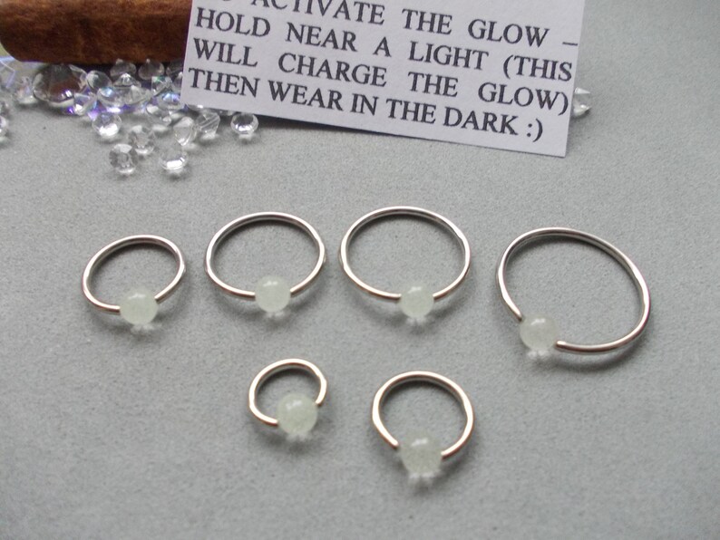 Puede incluir: Una colecci&oacute;n de aros para la nariz de color plateado con una peque&ntilde;a cuenta redonda y brillante. Los aros var&iacute;an en tama&ntilde;o y est&aacute;n dispuestos sobre una superficie gris. En el fondo se ve una peque&ntilde;a tarjeta con instrucciones.