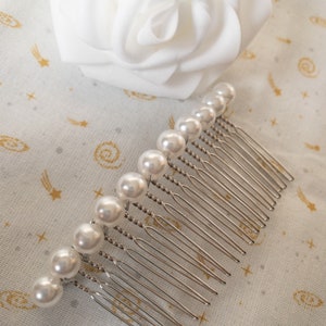 May include: A silver hair comb with a row of white pearl beads. The comb has a twisted design and is on a white fabric background with a gold star pattern.