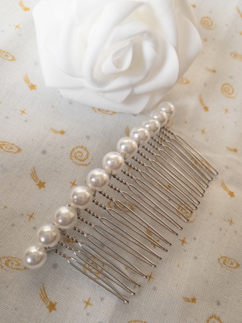 May include: A silver hair comb with a row of white pearl beads. The comb has a twisted design and is on a white fabric background with a gold star pattern.