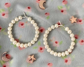 May include: A pair of pearl hoop earrings on a floral patterned fabric background. The earrings are made of white pearls and silver metal.
