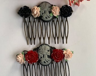May include: Two black metal hair combs with a cameo of a skull and flowers. The top comb has black flowers and the bottom comb has red flowers.