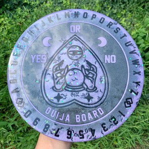 May include: A purple and black Ouija board with a heart-shaped planchette and a crescent moon on either side of the planchette. The board has the letters of the alphabet, numbers 0-9, and the words "yes", "no", and "goodbye".