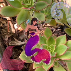 May include: A purple and pink mermaid keychain with a silver key ring. The mermaid is holding a small blue bird in her hand. The keychain is surrounded by green succulents.