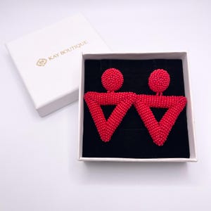 May include: A pair of red beaded earrings in a white box with the text "KAY BOUTIQUE". The earrings have a geometric design with a circular top and a triangular bottom. The box is open, revealing the earrings on a black velvet surface.