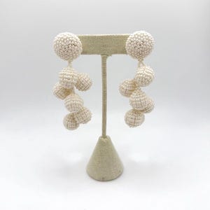 May include: A pair of off-white beaded earrings. Each earring features a large, round stud and a cascade of smaller, spherical beads. The earrings are displayed on a beige jewelry stand against a white background. The beads have a textured surface.