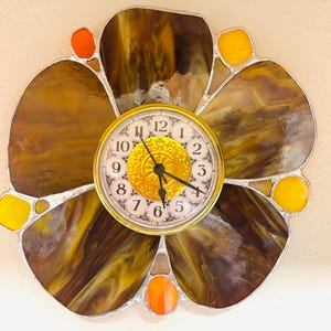 May include: A flower-shaped wall clock featuring large, swirled brown stained glass petals, accented by smaller orange and yellow glass pieces. The center is a round gold-rimmed clock face with black numbers and hands on a textured gold background.