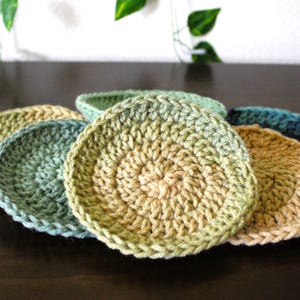May include: A collection of handmade, circular crochet coasters in various shades of green, blue, and tan. The coasters are arranged on a dark brown surface, showcasing their textured, spiral design. The yarn colors blend, creating a gradient effect.