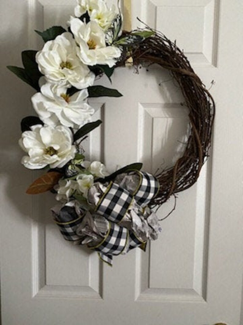 Peut inclure: Une couronne d&eacute;corative avec une base circulaire en vigne, orn&eacute;e de grandes fleurs de magnolia blanches et de feuilles vert fonc&eacute;. Un ruban &agrave; carreaux noir et blanc ajoute un accent contrastant. La couronne est con&ccedil;ue pour la d&eacute;coration de porte ou de mur.
