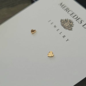 May include: Two gold heart-shaped earrings on a white card with the text "Mercedes L. Jewellery".