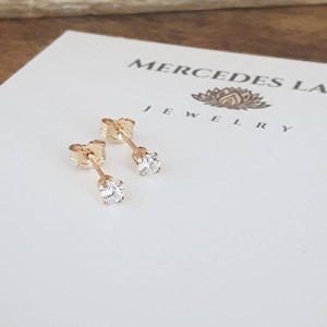 May include: A pair of gold stud earrings with small, clear round gemstones. The earrings are on a white card with the text "Mercedes La Jewellery" and a lotus flower design.