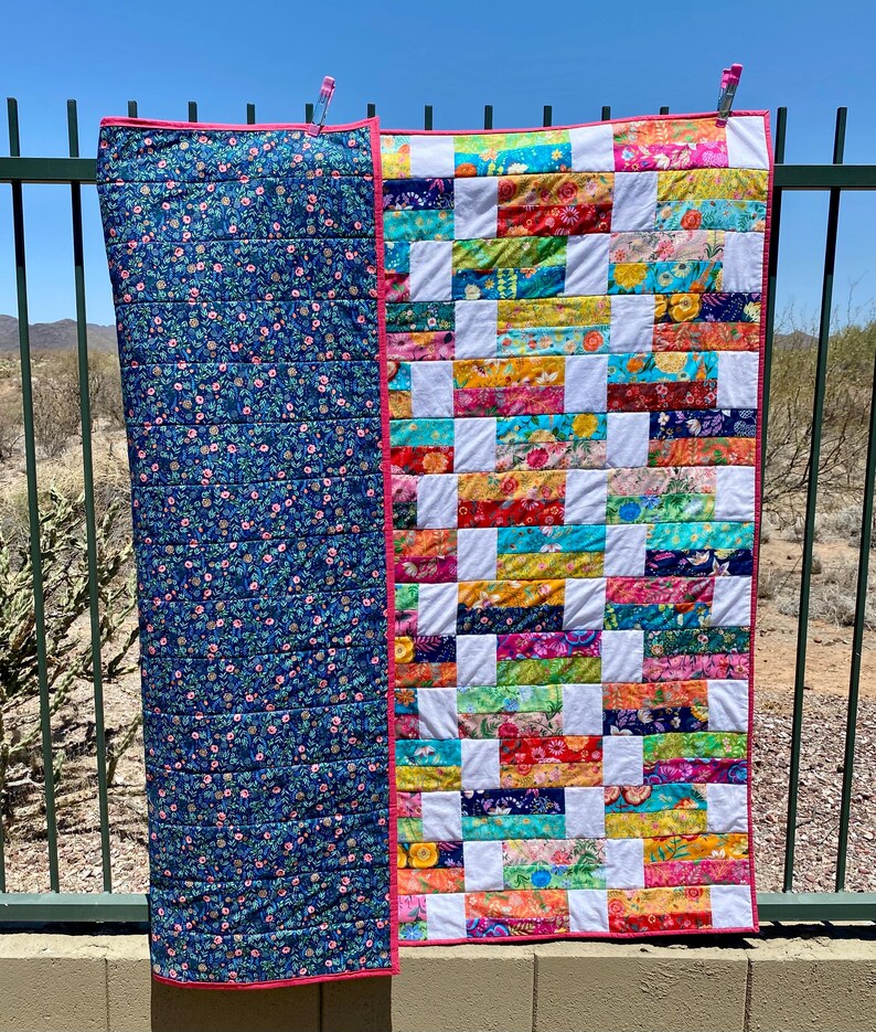 May include: A handmade quilt with a vibrant patchwork design and a floral patterned backing. The patchwork side features a colorful array of fabrics in various patterns and hues, with white squares separating the rectangles. The quilt is trimmed with a pink border.