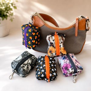 May include: A gray leather hobo bag with a brown strap, accompanied by five small, rectangular, zippered pouches featuring various Halloween-themed prints including candy corn, skulls, sunflowers, ghosts, and floral patterns with skulls.
